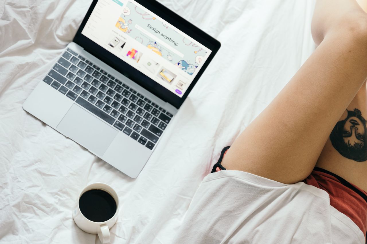 Vue plongeante d'une femme non reconnaissable, avec un tatouage sur la jambe, naviguant sur un netbook et buvant du café tout en se reposant sur un lit dans une chambre douillette